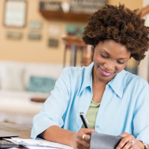 Pretty African American woman concentrates while writing a check or balancing her checkbook. Personal bills or documents are on the desk along with a laptop.